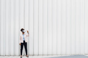 Businesswoman wearing virtual reality simulator gesturing in front of white wall - MEUF05459