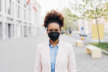 Afro businesswoman wearing protective face mask for smog standing on footpath - MEUF05470