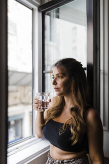 Junge Frau hält ein Glas Wasser und schaut durch ein Fenster in ihrem Haus - DCRF01158