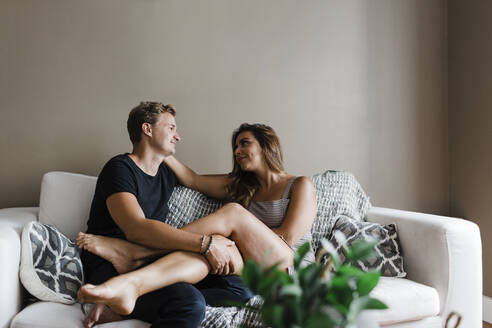 Lächelndes junges Paar, das sich gegenseitig ansieht, während es zu Hause auf dem Sofa sitzt - DCRF01169