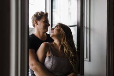 Smiling young couple looking at each other standing by window at home - DCRF01171