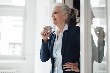 Glückliche Geschäftsfrau mit Kaffeetasse und Hand auf der Hüfte im Büro stehend - JOSEF08894