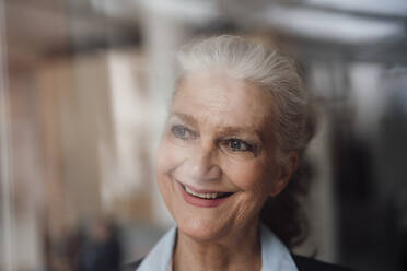 Happy senior businesswoman with gray hair looking through window - JOSEF08907