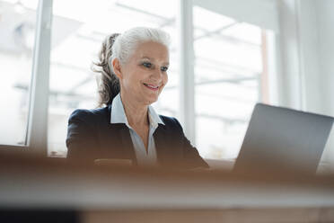 Happy senior businesswoman working on laptop at desk in office - JOSEF08909