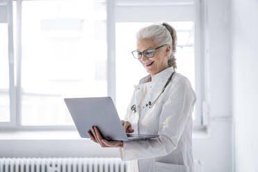 Happy senior doctor using laptop standing at the window - JOSEF08918
