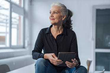 Senior businesswoman with tablet PC sitting on desk in office - JOSEF08933