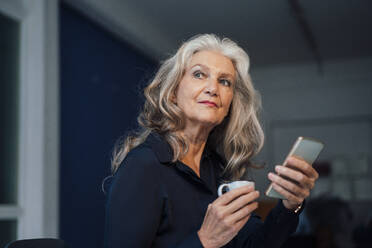 Senior businesswoman holding coffee cup and smart phone in office - JOSEF08947