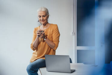 Smiling businesswoman with coffee cup sitting on desk at office - JOSEF08972