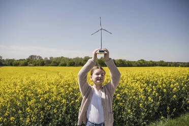 Lächelndes Mädchen mit geschlossenen Augen, das ein Modell einer Windkraftanlage auf einem Feld hält - UUF25866