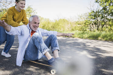 Lächelndes Mädchen schiebt Großvater, der auf einem Skateboard sitzt - UUF25881