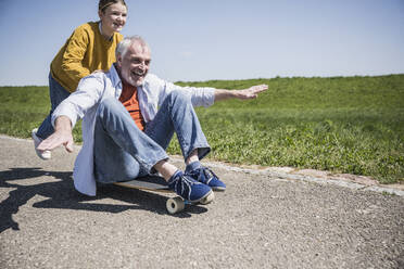 Glückliches Mädchen schiebt Großvater, der mit ausgestreckten Armen auf einem Skateboard sitzt - UUF25882