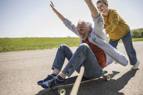 Playful girl pushing cheerful grandfather sitting with arms raised on skateboard - UUF25884
