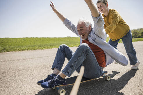 Verspieltes Mädchen schiebt fröhlichen Großvater, der mit erhobenen Armen auf einem Skateboard sitzt - UUF25884