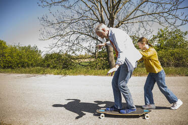 Verspieltes Mädchen, das seinen Großvater auf dem Skateboard schiebt - UUF25885