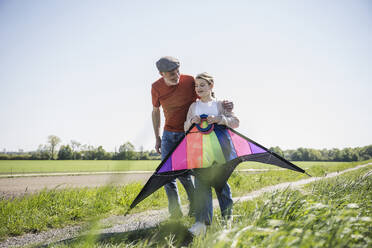 Grandfather walking with granddaughter holding kite walking on field - UUF25895