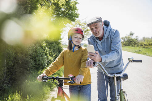 Großvater zeigt seiner Enkelin, die mit dem Fahrrad auf der Straße steht, das Mobiltelefon - UUF25896