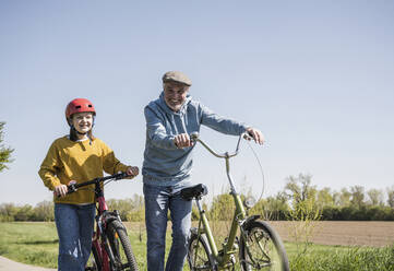 Happy grandfather and granddaughter walking with bicycle on sunny day - UUF25900