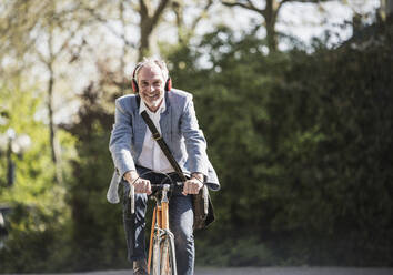 Smiling businessman listening music through wireless headphones cycling on sunny day - UUF25908