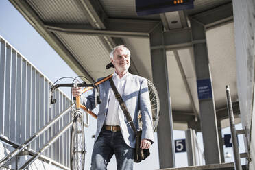 Smiling businessman carrying bicycle on shoulder walking at railroad station - UUF25911