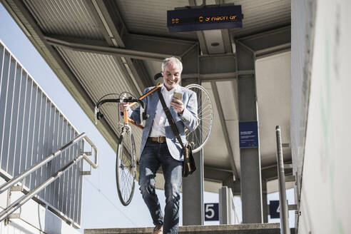 Glücklicher Geschäftsmann, der ein Fahrrad auf der Schulter trägt und ein Mobiltelefon benutzt, während er von einer Treppe heruntergeht - UUF25912