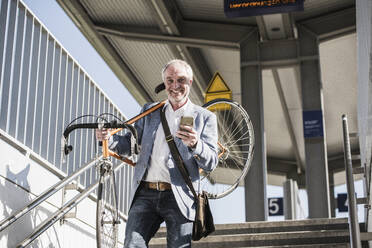 Lächelnder Geschäftsmann, der ein Fahrrad auf der Schulter trägt und ein Mobiltelefon in der Hand hält, als er die Treppe hinuntergeht - UUF25914