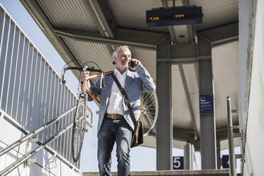 Smiling businessman carrying bicycle on shoulder talking through smart phone at railroad station - UUF25915