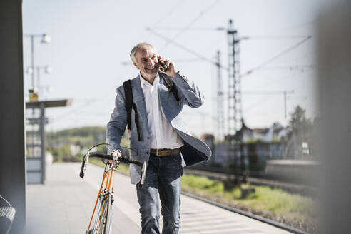 Glücklicher Geschäftsmann, der mit dem Handy telefoniert und mit dem Fahrrad am Bahnsteig spazieren geht - UUF25917