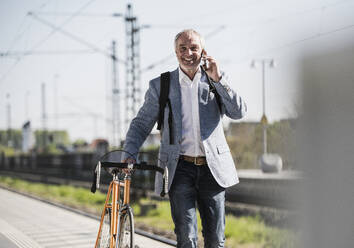 Smiling businessman talking on mobile phone walking with bicycle at railroad station platform - UUF25918