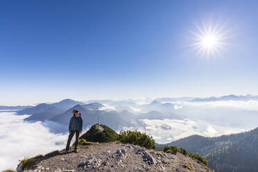 Deutschland, Bayern, Sonne scheint über einer Wanderin auf einem Berggipfel mit dichtem Nebel und dem Gipfel des Herzogstands im Hintergrund - FOF13153