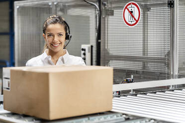 Happy businesswoman wearing headset with box on conveyor belt at warehouse - PESF03699