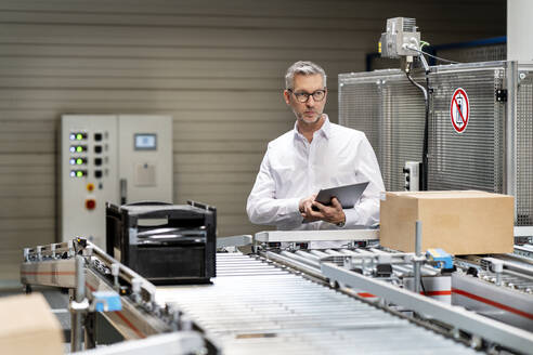 Älterer Geschäftsmann, der einen Karton auf einem Förderband mit einem Tablet-PC in einer Fabrik untersucht - PESF03700