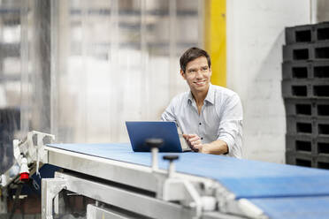 Lächelnder Geschäftsmann mit Laptop auf einem Förderband in einem Lagerhaus - PESF03724