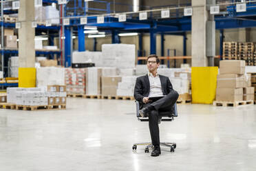 Confident businessman sitting on chair at warehouse - PESF03774