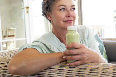 Smiling mature woman with bottle of smoothie sitting on sofa at home - ESTF00034