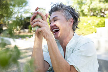 Cheerful woman with bottle of smoothie sitting in garden - ESTF00043