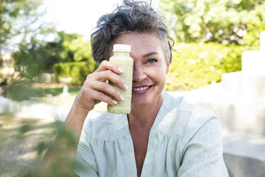 Lächelnde Frau, die ihr Gesicht mit einer Flasche Smoothie bedeckt, sitzt im Garten - ESTF00044