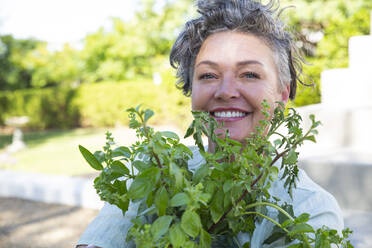 Lächelnde reife Frau mit Pflanze im Garten sitzend an einem sonnigen Tag - ESTF00046