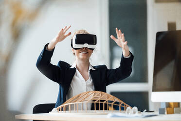 Businesswoman wearing virtual reality headset gesturing at office - JOSEF08983