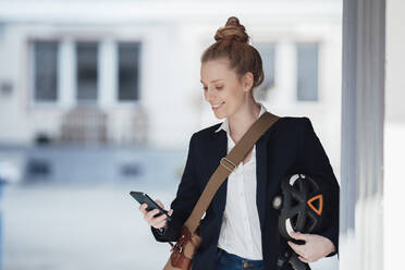 Lächelnde Geschäftsfrau mit Helm und Smartphone an der Wand - JOSEF08984