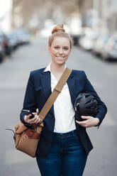 Happy businesswoman walking at street holding helmet and smart phone - JOSEF08991
