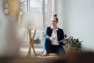 Contemplating businesswoman with tablet PC sitting near window at office - JOSEF09019