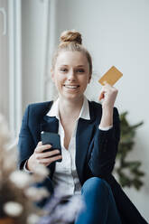 Smiling businesswoman holding credit card and smart phone at office - JOSEF09026