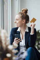 Contemplating businesswoman holding smart phone and credit card at office - JOSEF09027