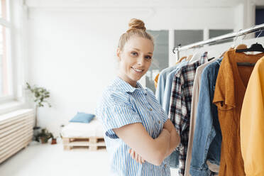 Lächelnde Modedesignerin mit verschränkten Armen, die in der Nähe eines Kleiderständers im Studio steht - JOSEF09037