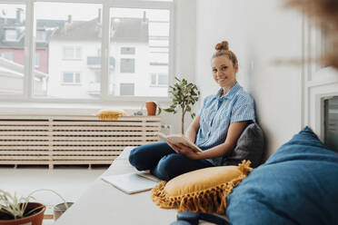 Smiling young freelancer with book sitting on bed at home - JOSEF09053