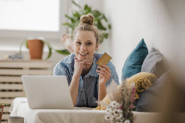 Smiling businesswoman with laptop and credit card lying on bed at home - JOSEF09055