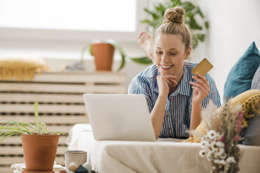 Smiling freelancer holding credit card looking at laptop lying on bed at home - JOSEF09056