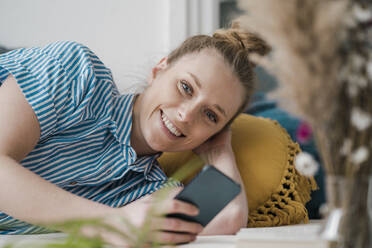 Lächelnder junger Freiberufler mit Smartphone, der zu Hause auf dem Bett liegt - JOSEF09068