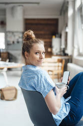 Businesswoman with drink and smart phone sitting on chair at home - JOSEF09080