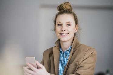 Lächelnde Geschäftsfrau mit Blazer und Smartphone im Büro - JOSEF09089